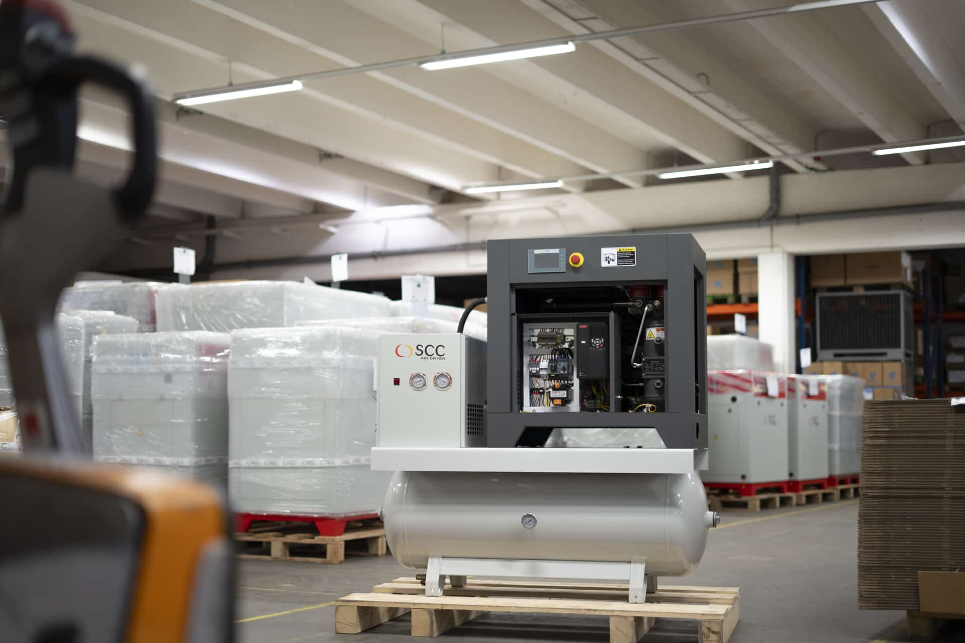 Industrial air compressor machine with control panel and gauges in a warehouse, surrounded by packaged equipment.